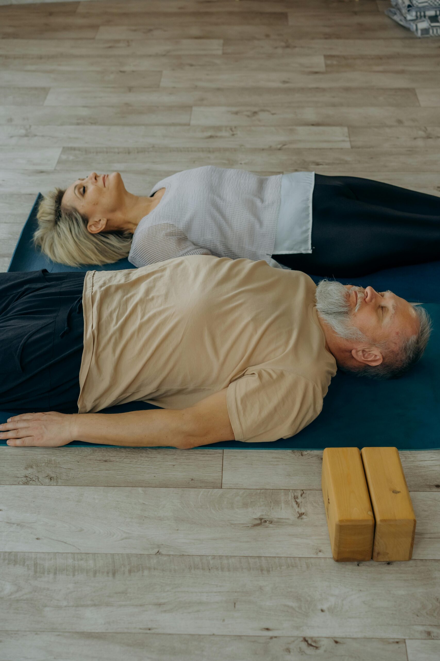 Elderly man and woman performing relaxation yoga on mats indoors with wooden blocks nearby.