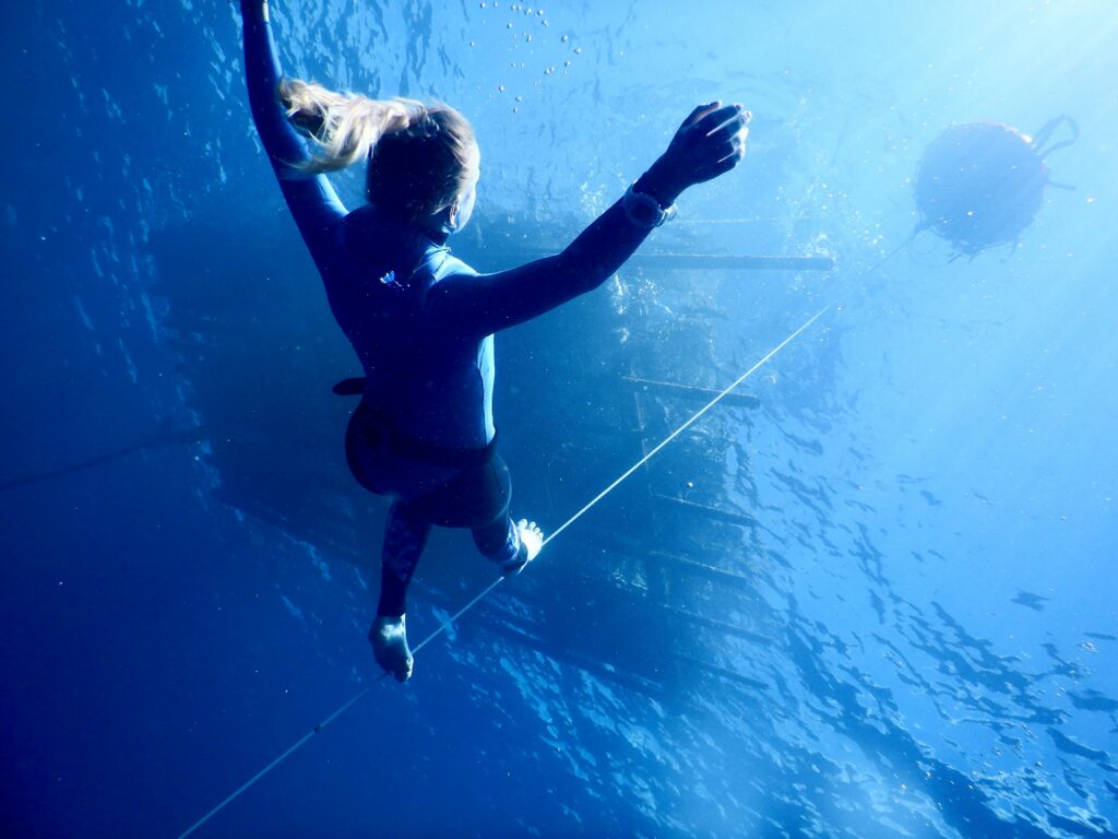 From below of unrecognizable young female diver with long hair in wetsuit swimming under transparent blue seawater on sunny day